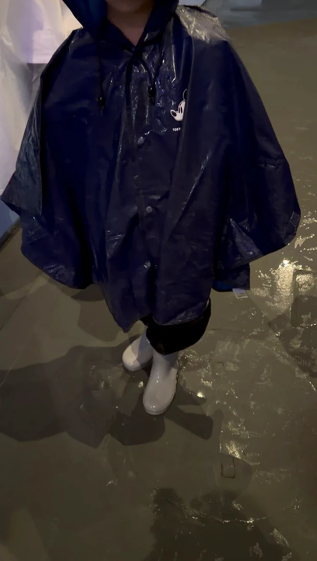 雨のディズニーランドで、黒いレインポンチョと白い長靴を身につけた子ども。水たまりに雨が反射し、雨の日ならではのパークの様子が伝わる一枚。